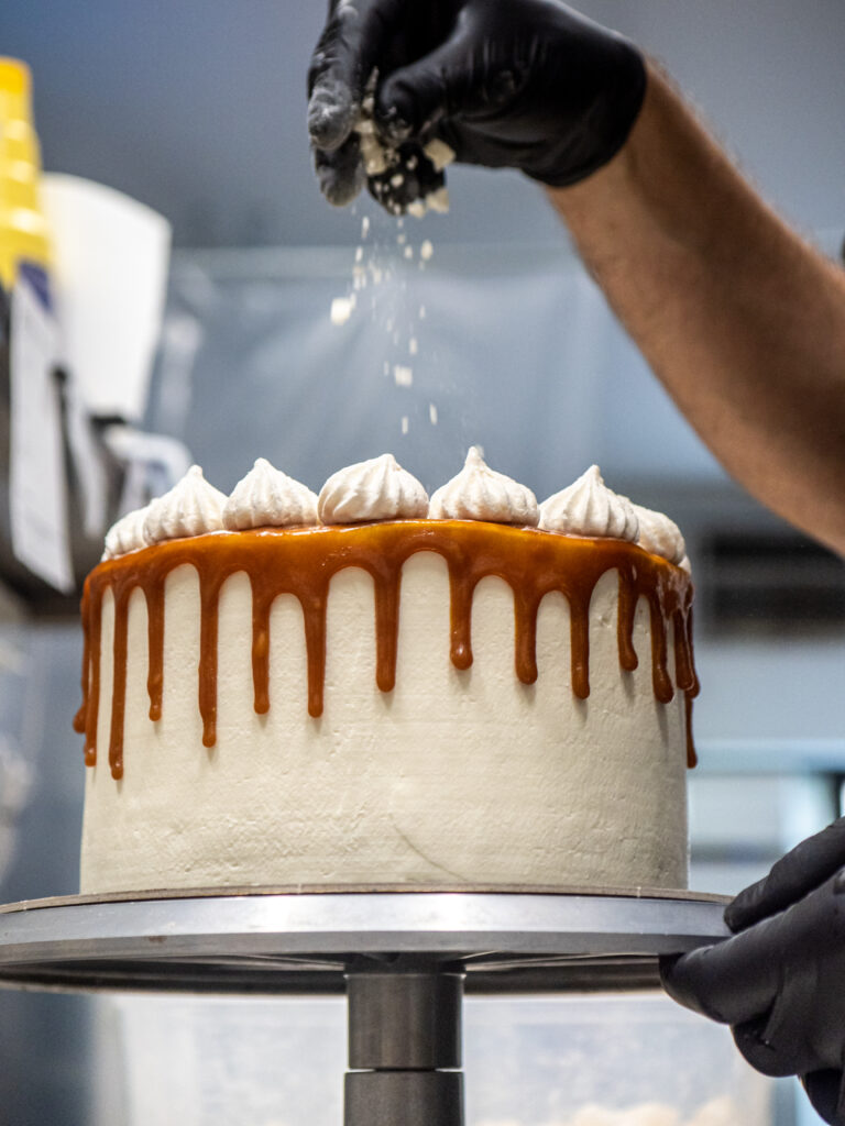 cake chef hand sprinkling break meringue on a frosted salty caramel dripping cake